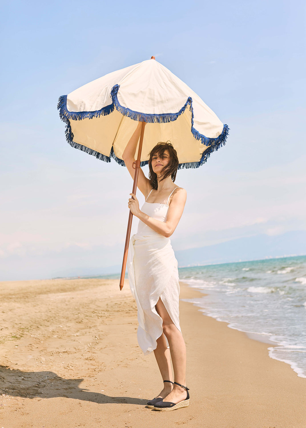 Woman walking along the beach wearing black espadrille sandals, holding a fringed beach umbrella in a Mediterranean coastal setting.