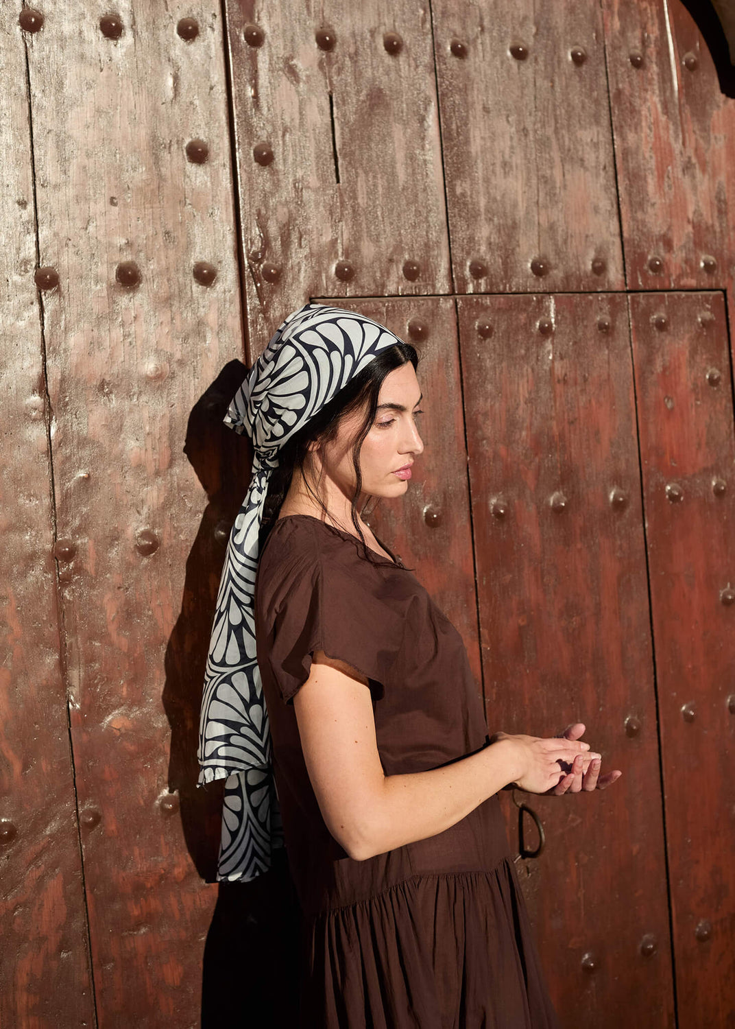 Woman standing against a rustic wooden door, wearing a flowing brown dress and the Foulard Scarf in Blue Palms. A quiet moment where light and shadow meet, evoking timeless Mediterranean charm.