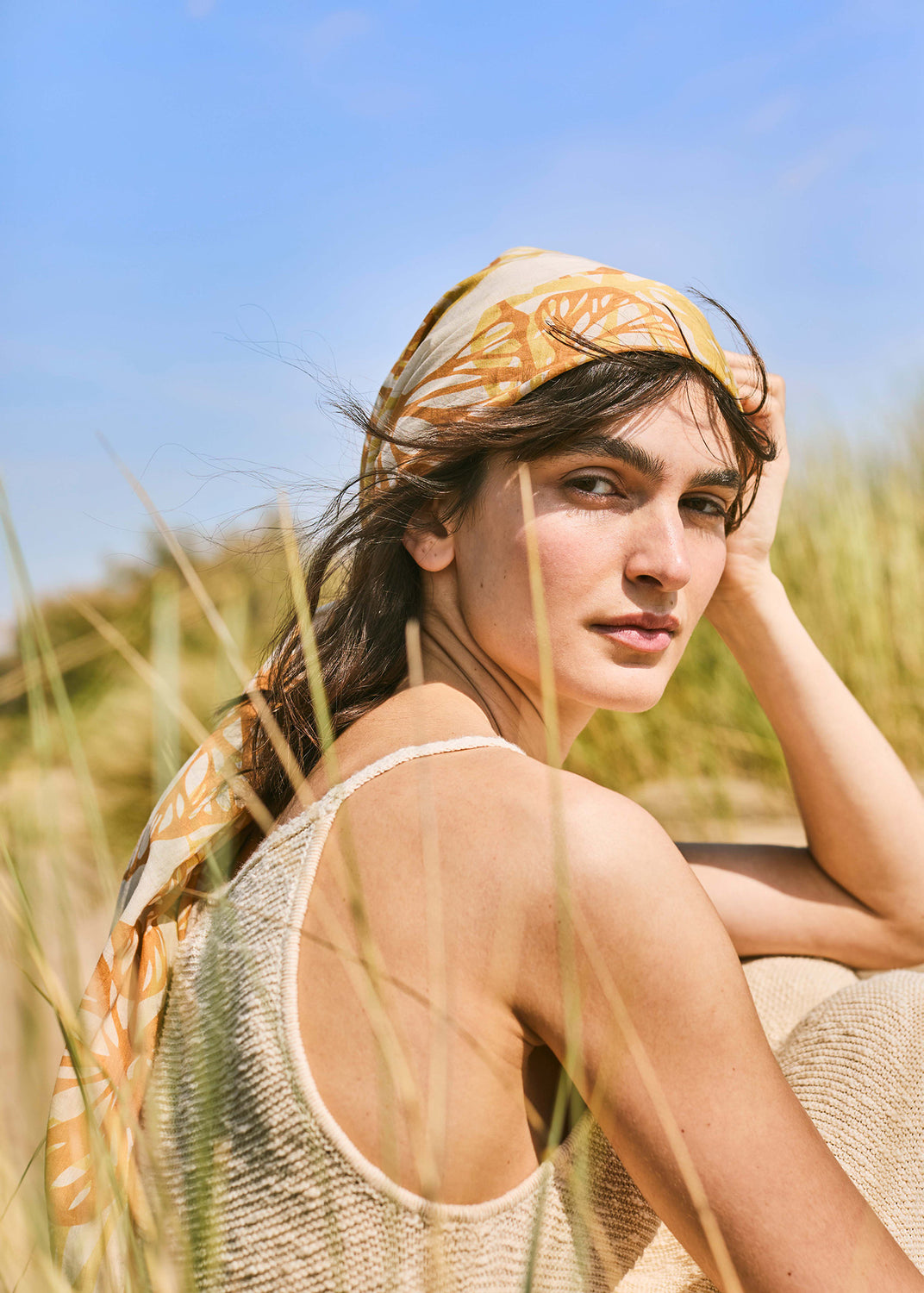 Woman wearing an orange floral scarf and lightweight knit top, styled among Mediterranean sand dunes in warm summer light.
