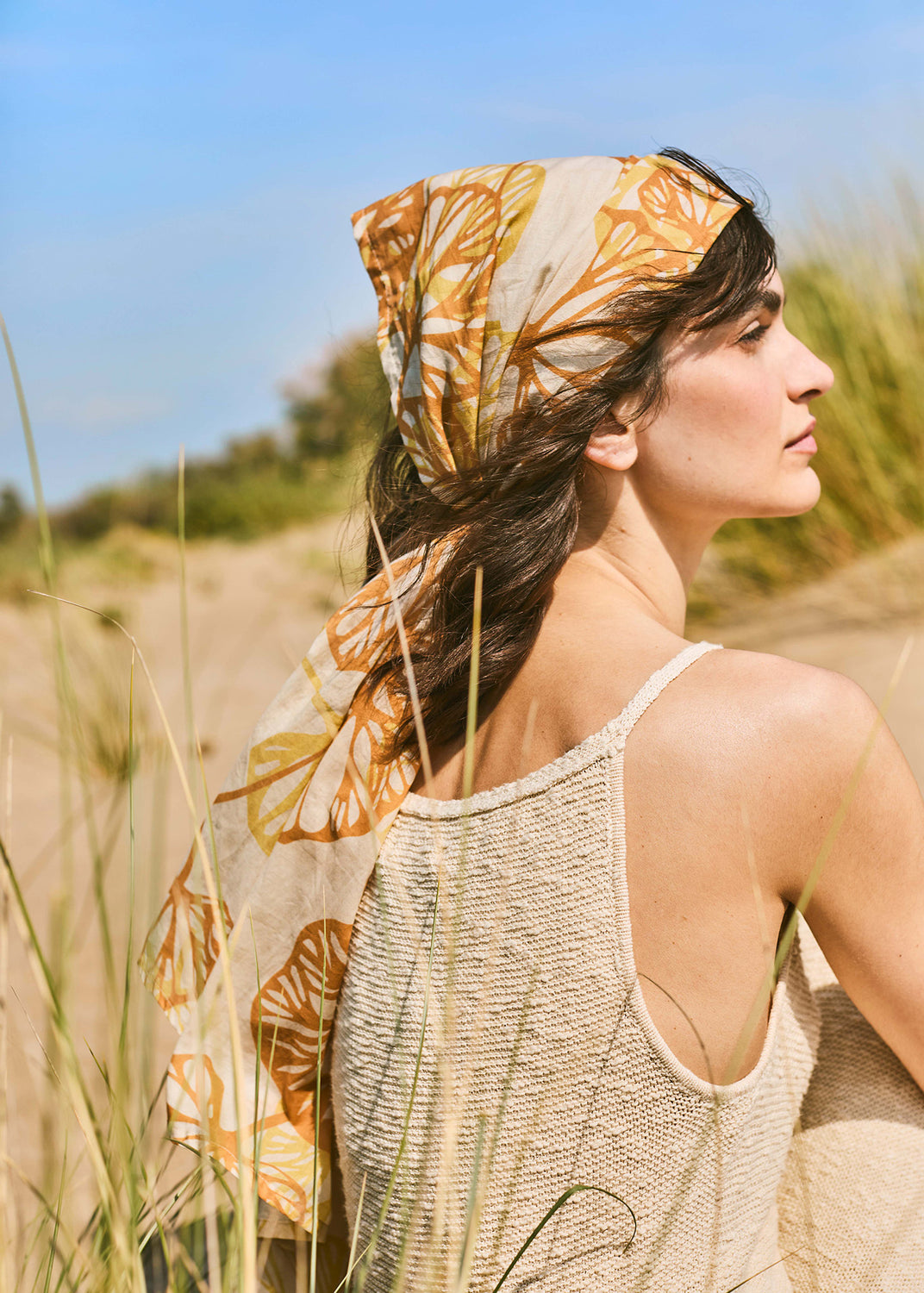Woman wearing a lightweight striped knit top and floral scarf standing among Mediterranean sand dunes in warm natural light.