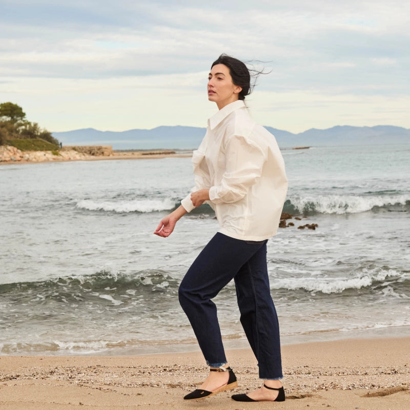 A woman walks along a sandy beach wearing a white oversized long-sleeve blouse, dark navy cropped pants, and black flats, with ocean waves and distant mountains in the background.