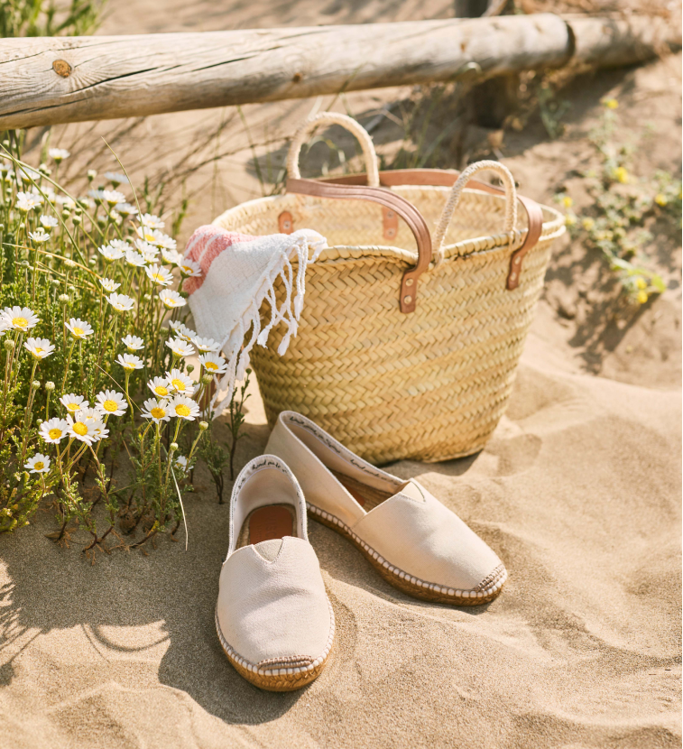Pair of beige flat espadrilles on sand with a straw bag and flowers in the background
