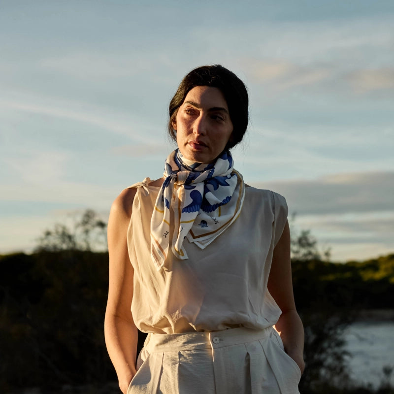 A woman wearing a cream sleeveless top and matching skirt, styled with a blue and white patterned silk scarf, standing outdoors against a soft sky background.