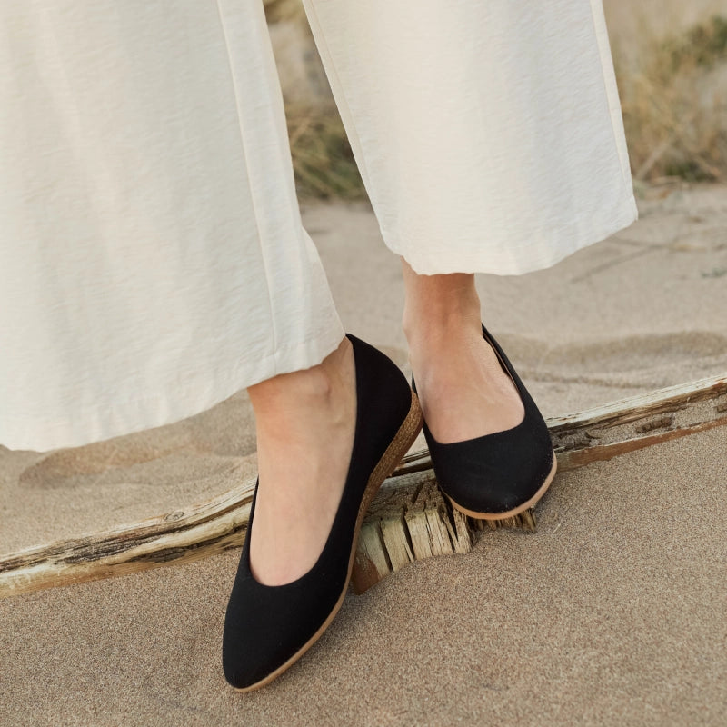 A person wearing black pointed-toe flats with a natural jute-wrapped heel, standing on sand next to a piece of driftwood.