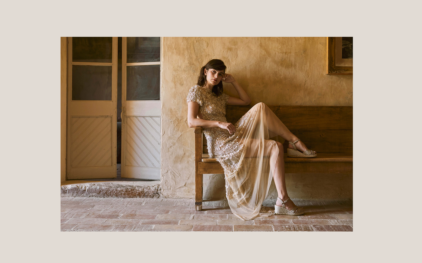 Woman wearing metallic champagne espadrille wedges and an embellished evening dress seated on a rustic wooden bench in a warm Mediterranean interior.
