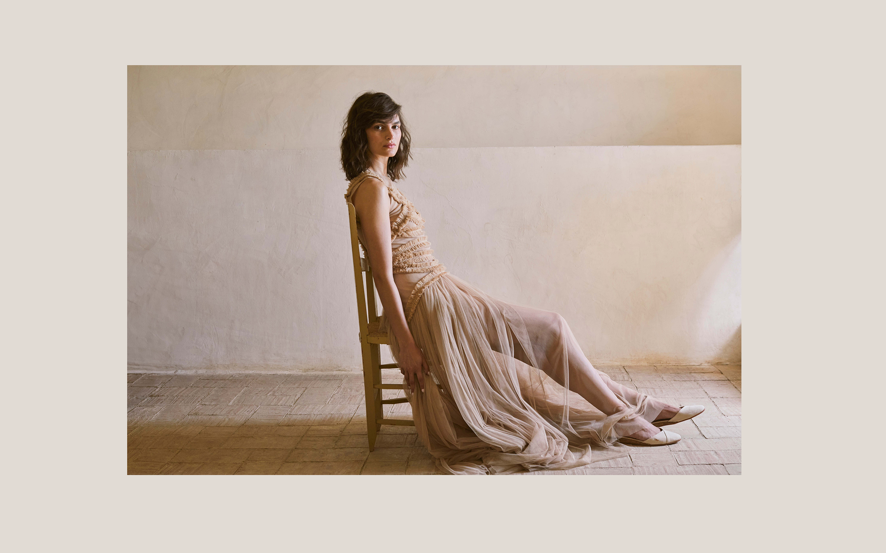 Woman wearing ivory beige espadrille flats and a flowing blush tulle dress seated in a softly lit Mediterranean interior.
