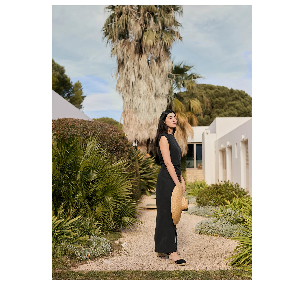 A woman stands on a gravel path in a garden courtyard, wearing a sleeveless black maxi dress and holding a wide-brimmed straw hat. White buildings, lush green plants, and a tall palm tree are visible in the background.