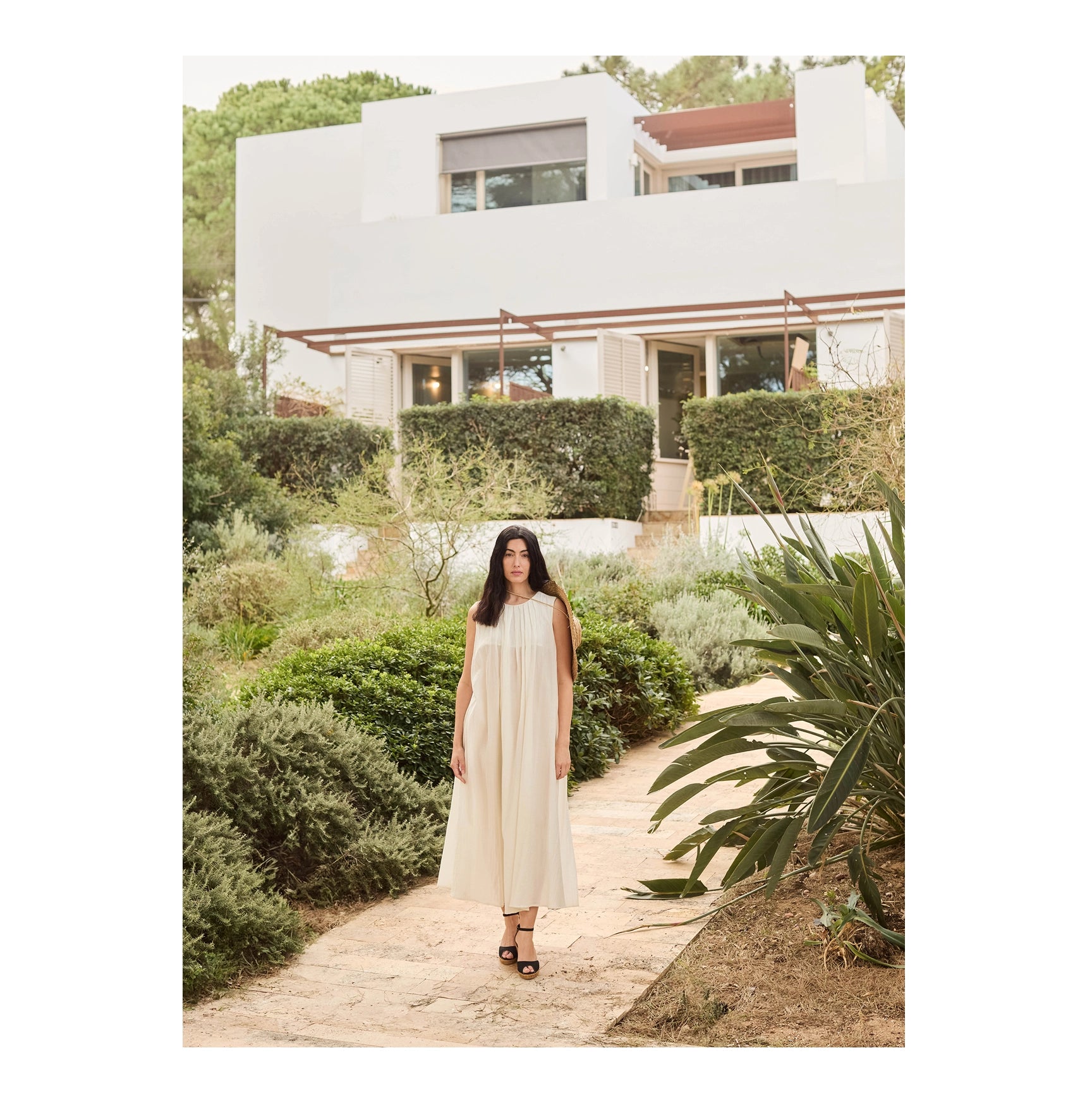 A woman wearing a sleeveless cream-colored midi dress and black sandals stands on a stone path in a garden, with a modern white house in the background.