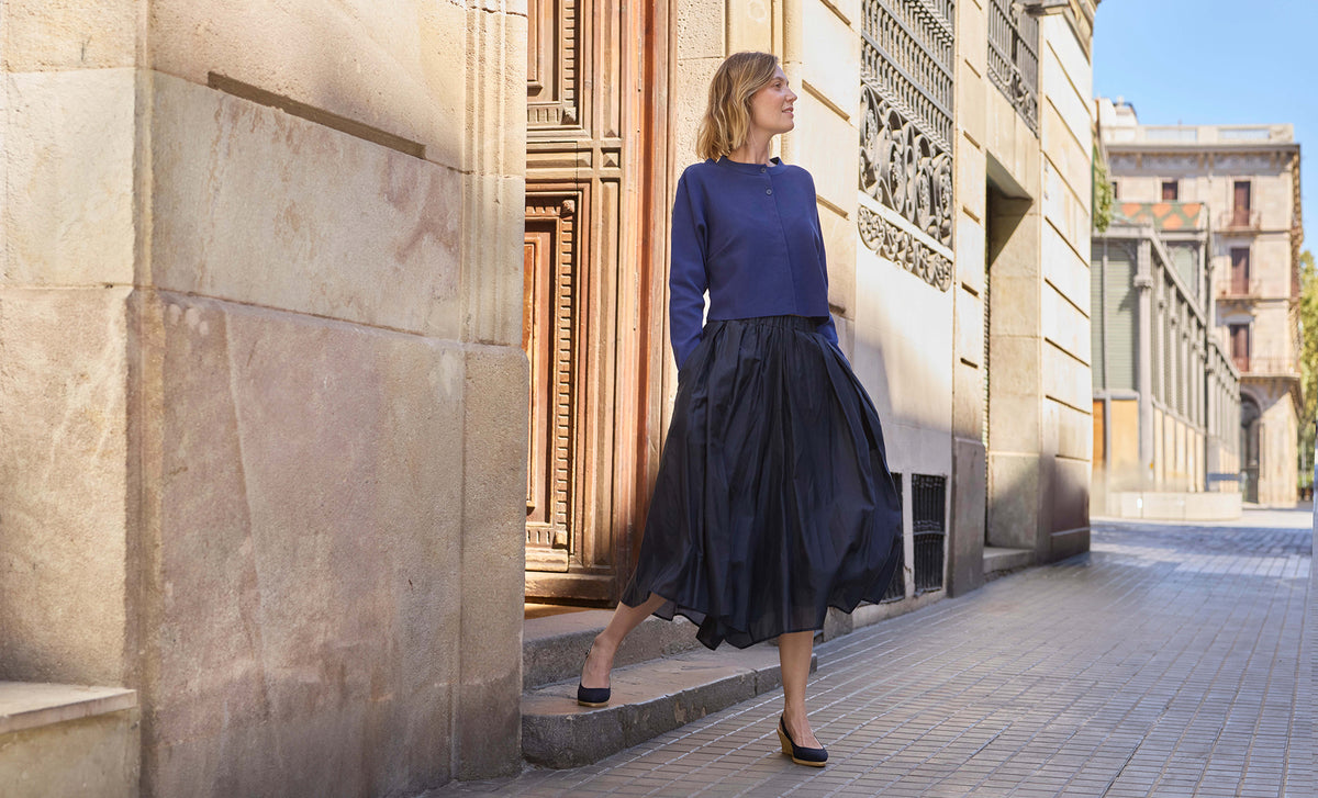 A woman stands on stone steps in an outdoor urban setting, wearing a navy blue long-sleeve top and a dark navy pleated midi skirt with black wedges.
