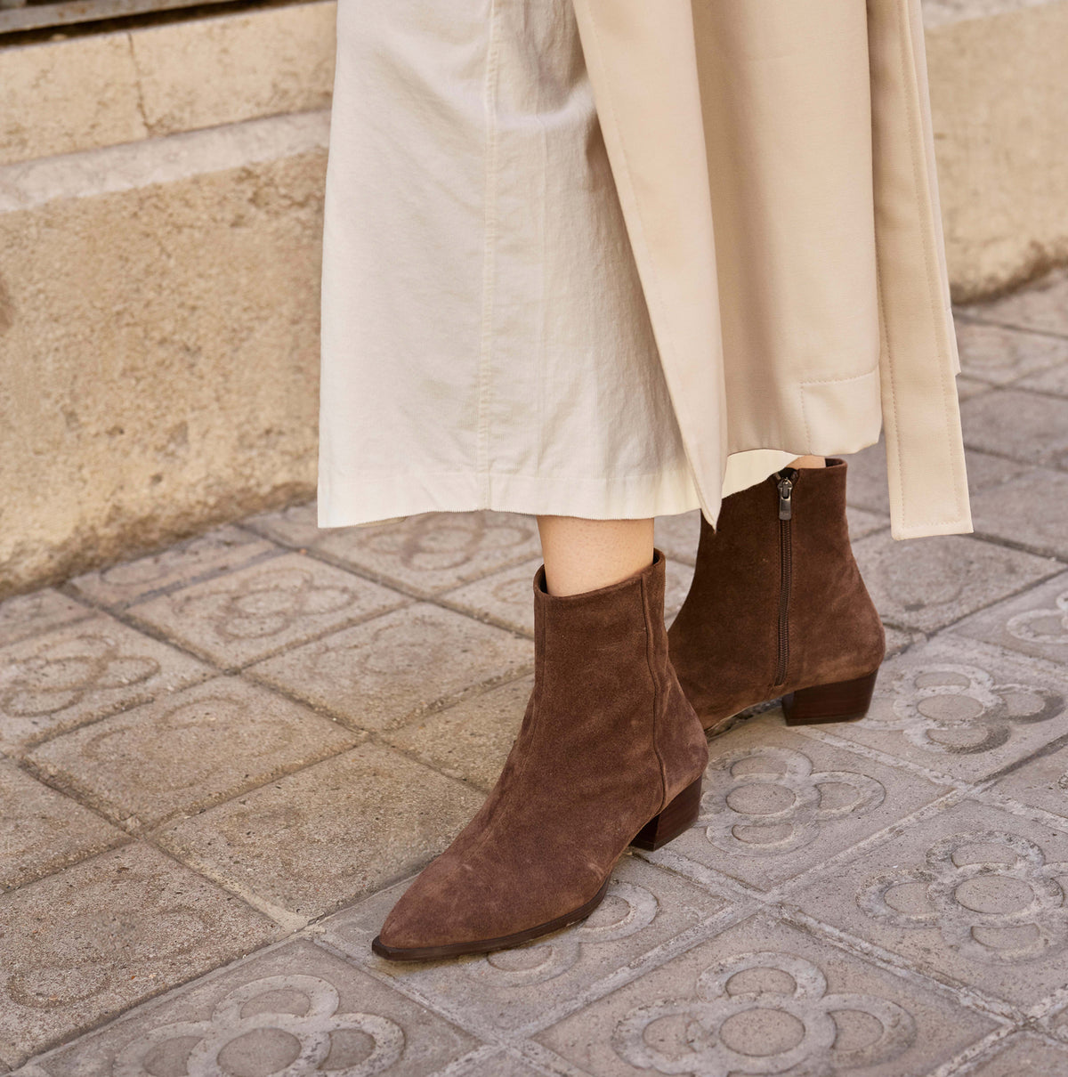A person wearing brown suede ankle boots with low block heels and side zippers, paired with a beige midi skirt, standing on a patterned stone sidewalk.