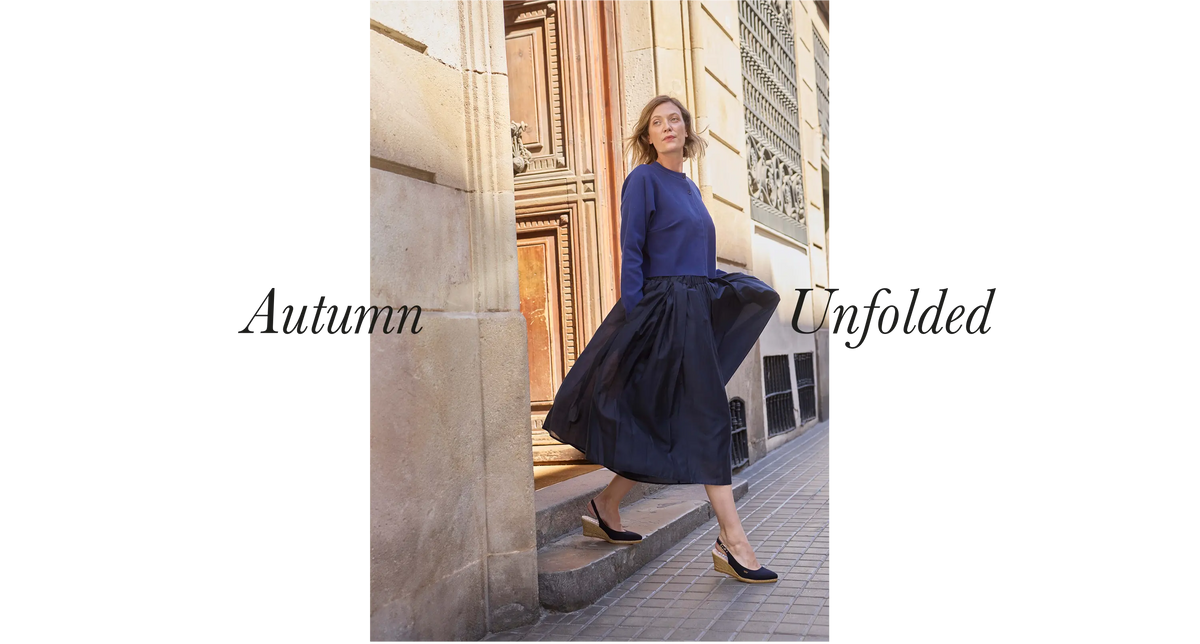 A woman stands on stone steps outside a building wearing a blue long-sleeve top and a voluminous dark navy skirt, with the text 'Autumn Unfolded' overlaid on both sides.