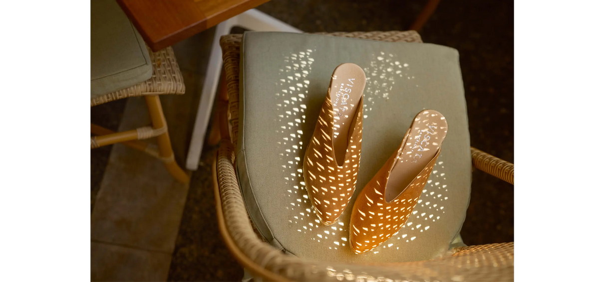 A pair of camel suede mule clogs with geometric cutout patterns, displayed on a sage green cushion inside a wicker chair.