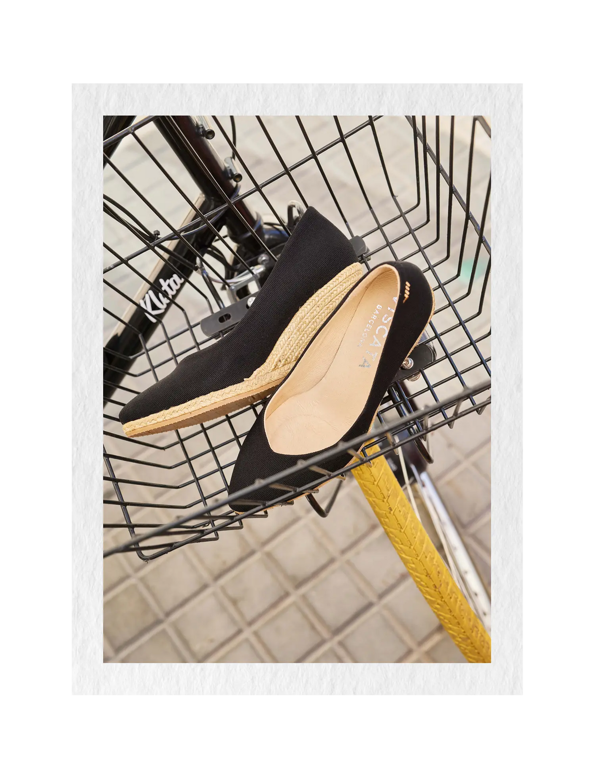 A pair of black pointed-toe wedes with espadrille-style jute soles, displayed on a bicycle basket over a tiled floor.