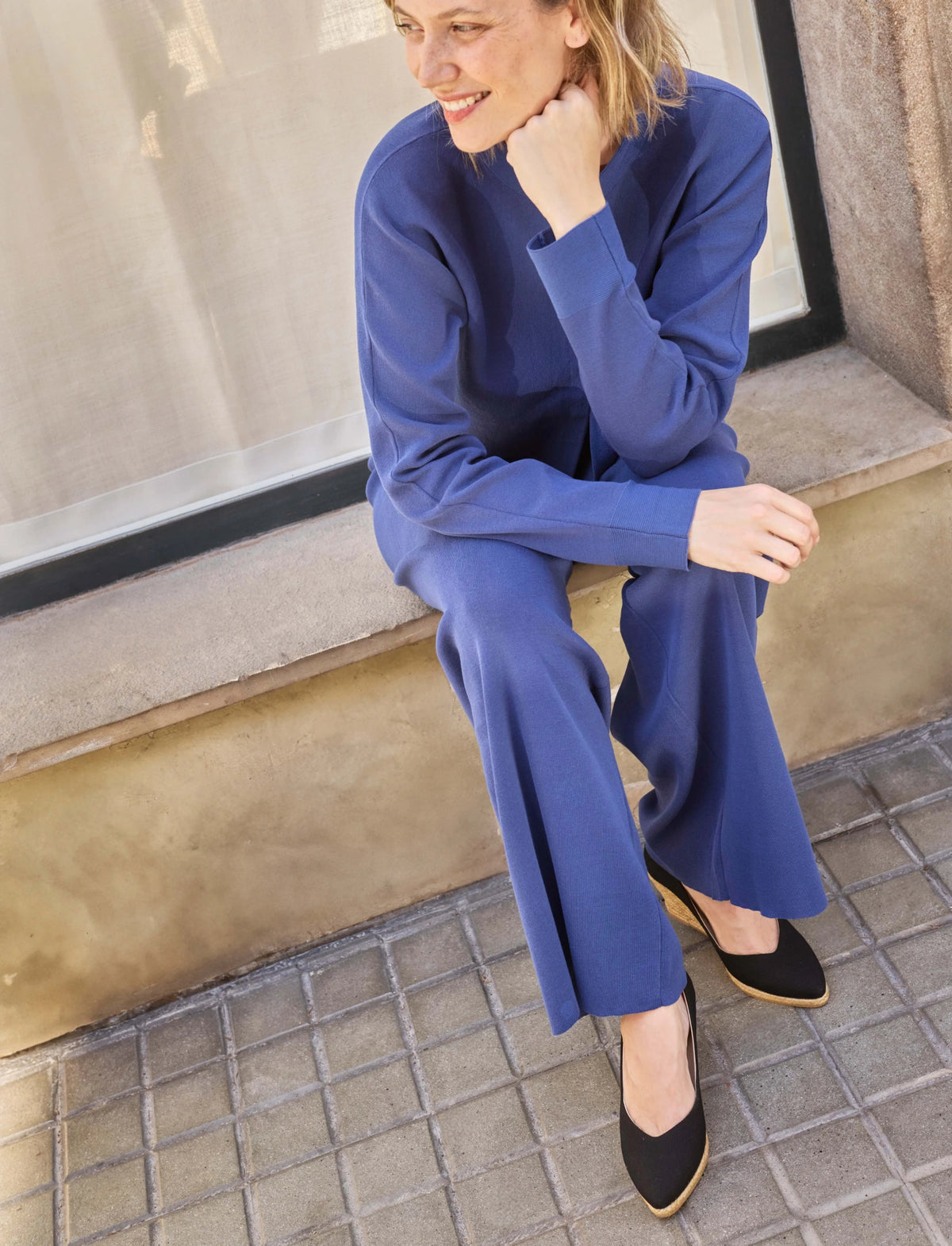 A woman wearing a matching royal blue long-sleeve top and wide-leg trousers sits on a stone ledge next to a window, wearing black wedge espadrilles.