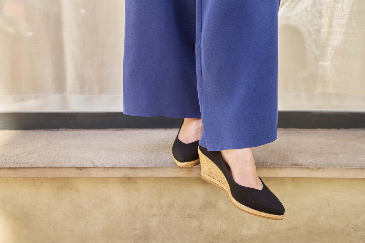 A person wearing blue wide-leg trousers and black wedge espadrilles, standing on a beige stone ledge.