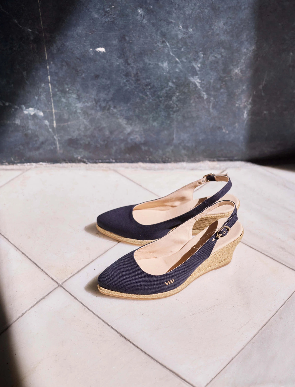 A pair of navy blue wedge slingback shoes with espadrille-style jute-wrapped heels, displayed on a light tiled floor against a dark textured wall.