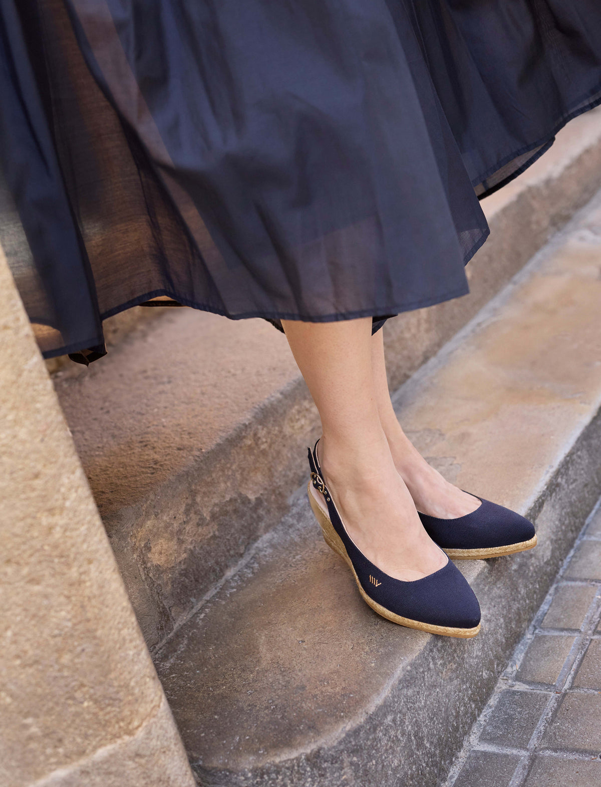 A person wearing navy blue slingback wedge espadrilles with a sheer navy midi skirt, standing on stone steps.