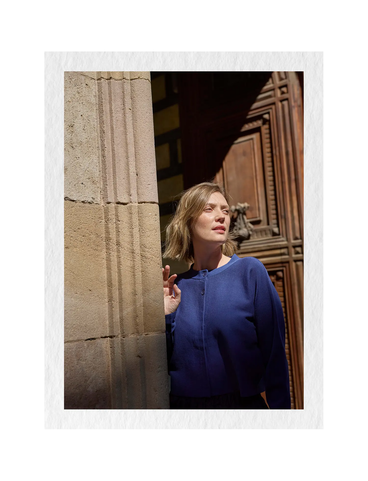 A woman wearing a blue knit top stands in a doorway next to a stone pillar, with a white textured border around the photo.