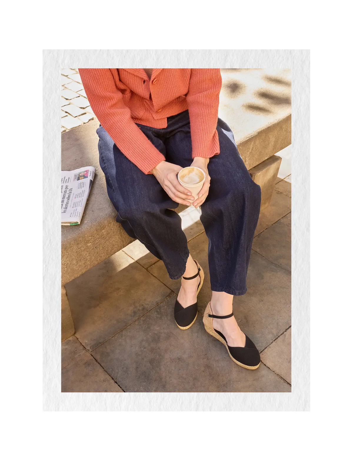 A person seated on a stone bench outdoors wearing dark navy wide-leg pants, a coral-orange ribbed long-sleeve top, and black low wedge espadrilles with tan wedge soles. A cup is held in the hands, with a newspaper on the bench beside them.