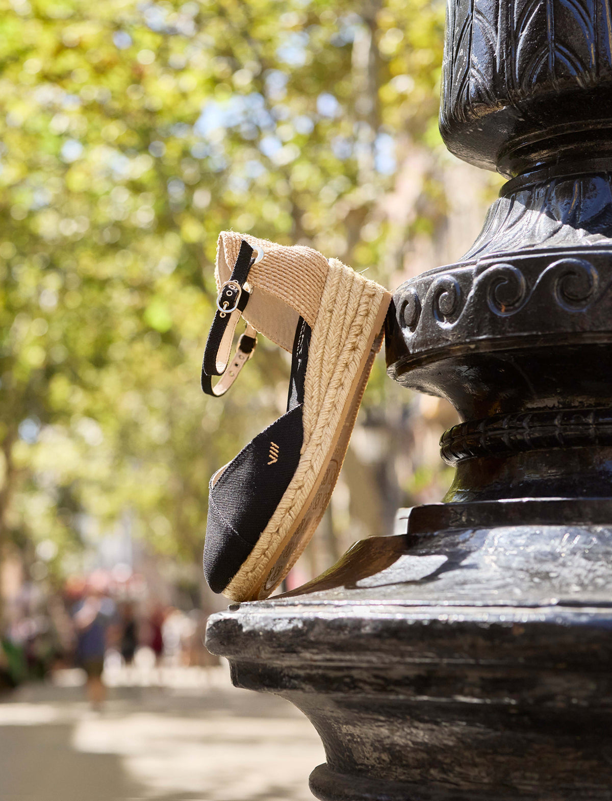 A pair of black low wedge espadrilles with ankle straps, photographed outdoors against a blurred park background with a dark metal column in the foreground.