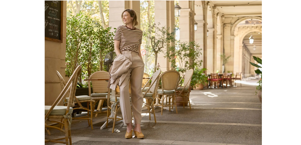 A woman stands in a sunlit indoor-outdoor corridor with columns, chairs, and plants, wearing a beige and brown striped short-sleeve top, beige trousers, and tan shoes, while holding a light beige jacket.