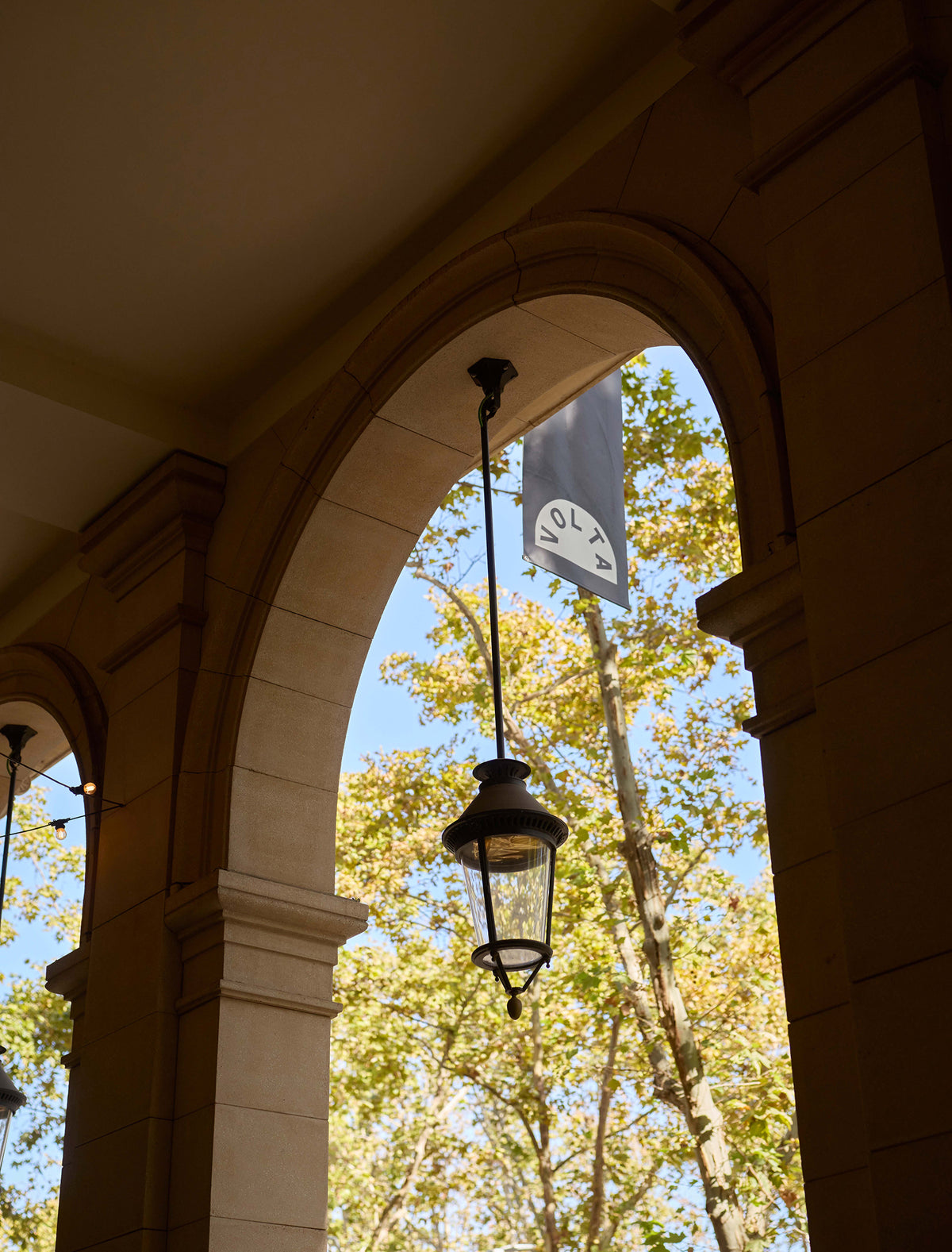 A hanging black lantern and a white banner with the circular 'VOLTA' logo are suspended under a large stone archway, with trees and blue sky visible in the background.
