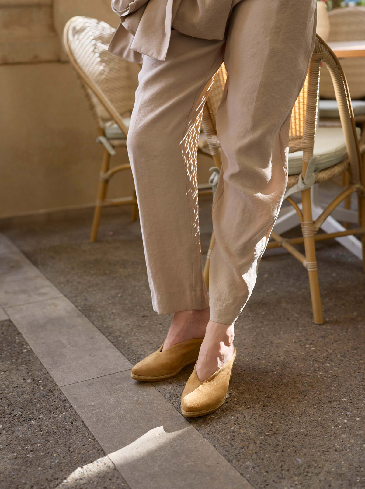 A person wearing loose beige trousers and camel slip-on shoes, standing on a stone patio with wicker chairs in the background.