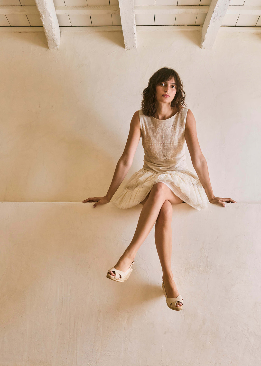 Woman wearing beige canvas slingback espadrille wedges with crossover straps, styled with an ivory dress in a warm Mediterranean interior.
