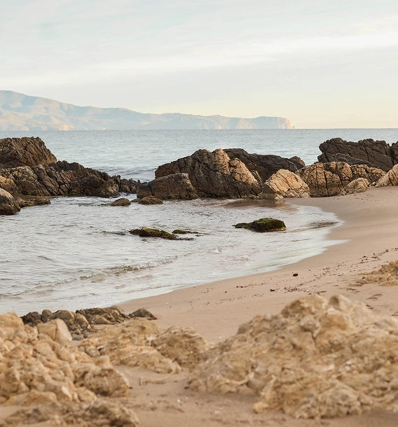 A scenic coastal landscape with sandy beach, rocky shoreline, calm water, distant mountains, and a pale sky.