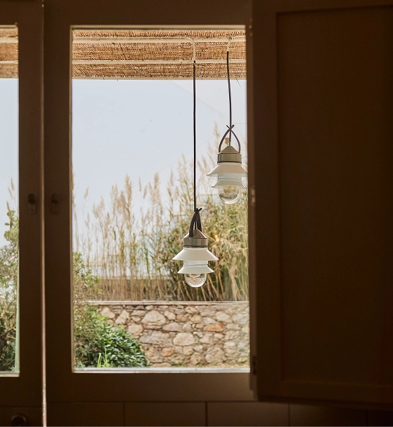 Two hanging pendant lights with white glass shades and dark metal frames, suspended in front of an open window looking out onto a reed field.