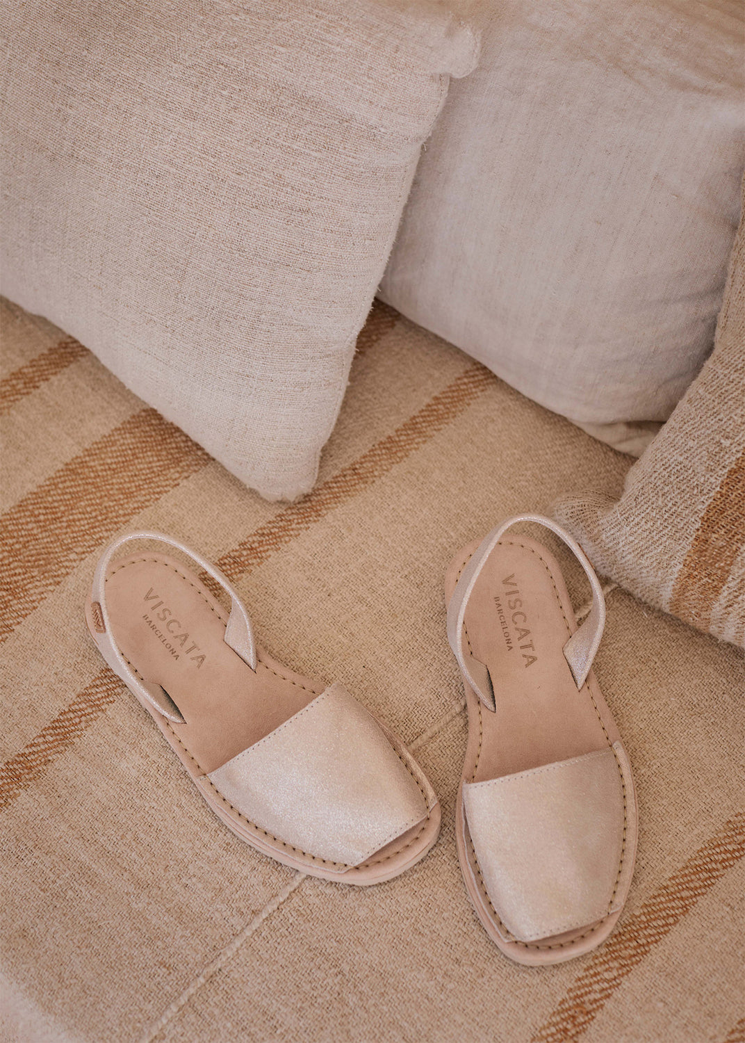 Two gold leather slingback menorquina flats lie on a striped woven cushion beside light linen pillows.