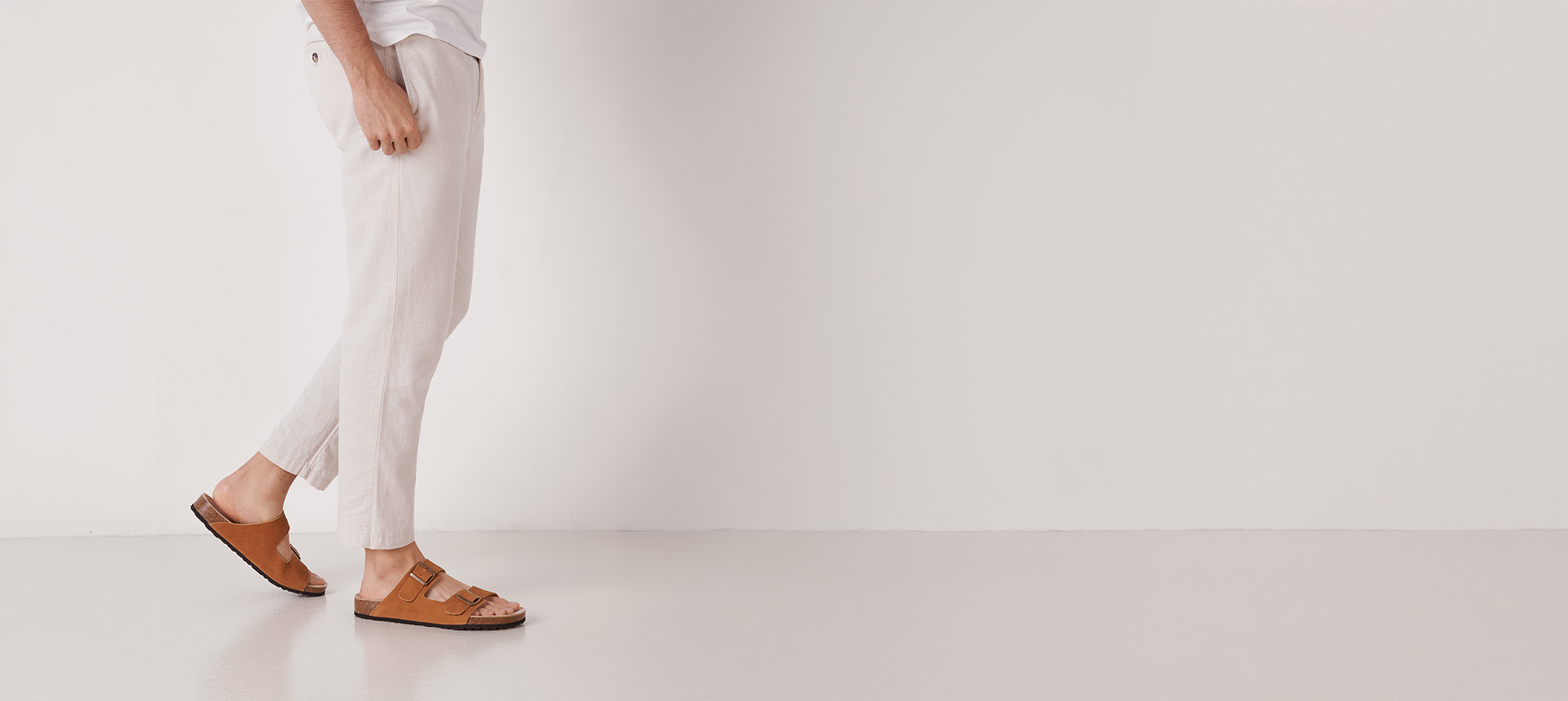 Person wearing tan suede double-buckle sandals styled with neutral linen trousers on a clean background.