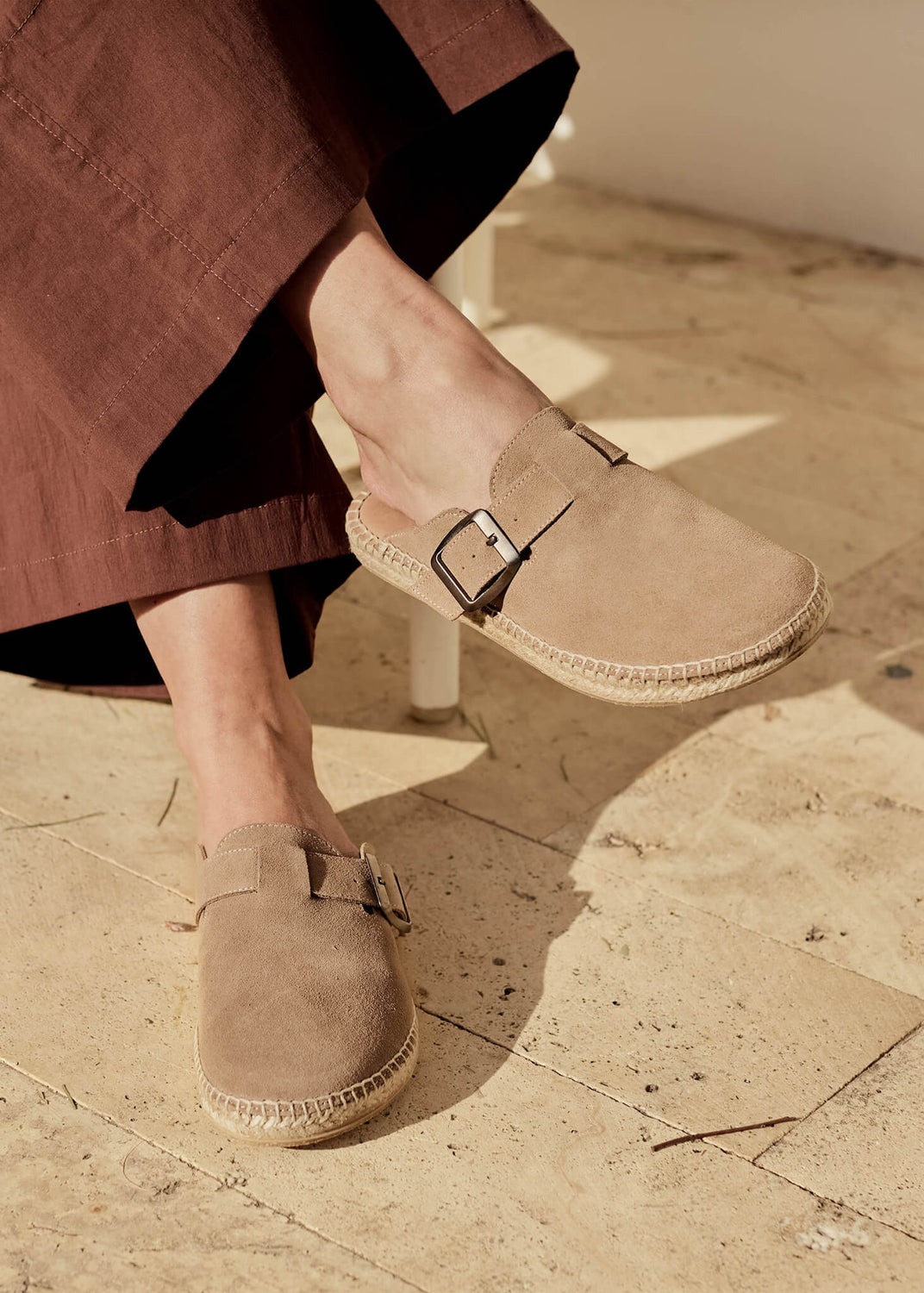 Close-up of a woman seated in the sun wearing Menuda camel suede slip-on flats with buckle detail, paired with flowing brown trousers. The warm tones and natural textures evoke relaxed Mediterranean simplicity.