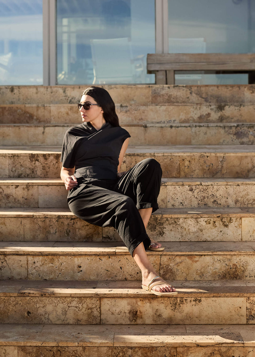Woman sitting on sunlit stone steps wearing Miramar camel suede strap flats and a relaxed black outfit, embodying understated Mediterranean ease and timeless elegance.