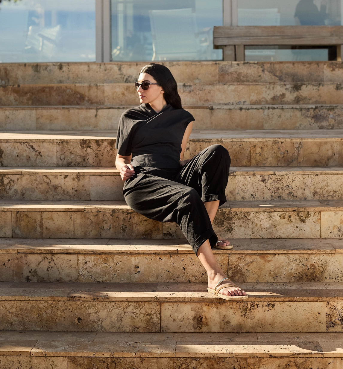 A woman wearing a black short-sleeve top and black cropped pants sits on stone steps outdoors, wearing beige sandals and dark sunglasses.