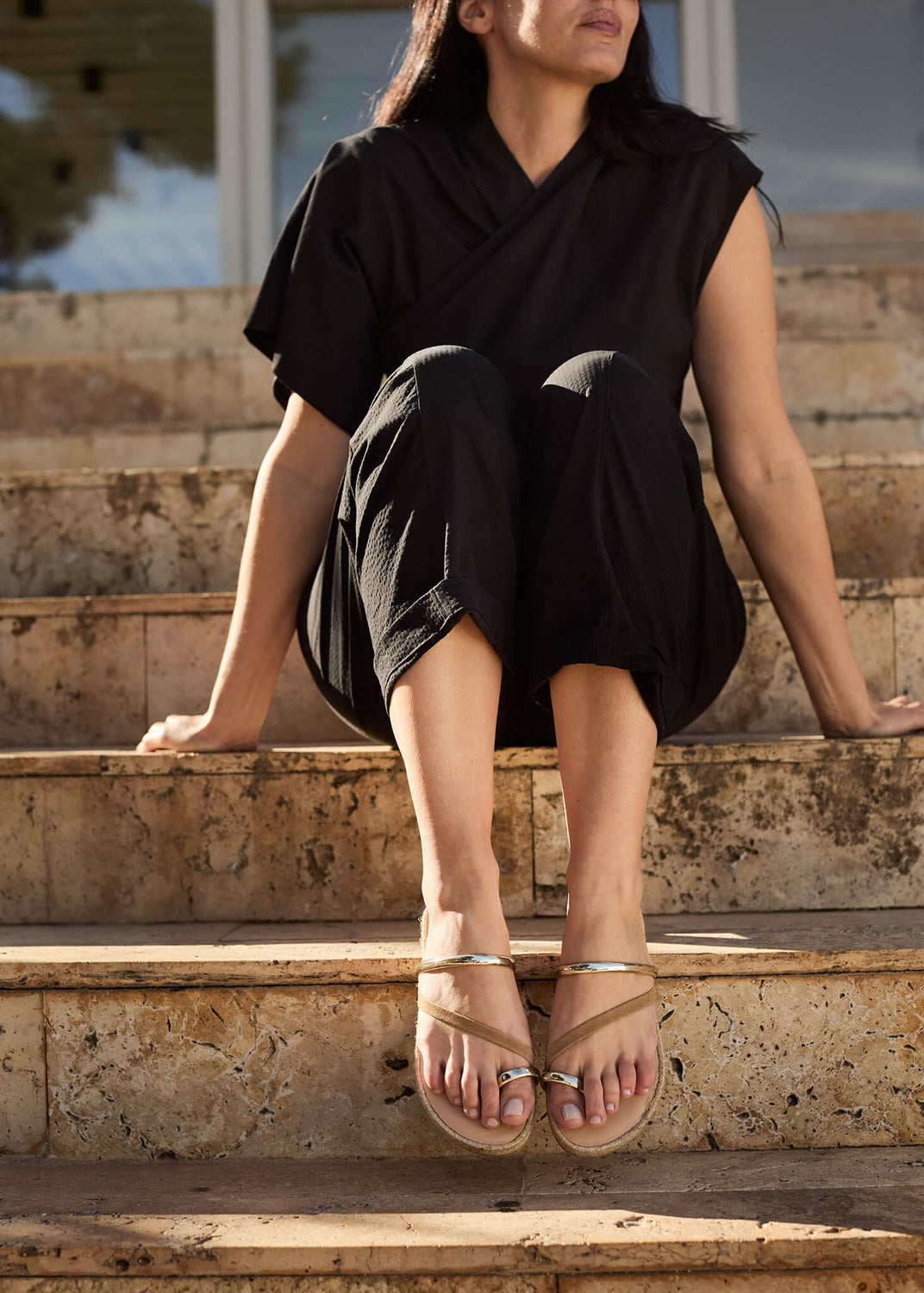 Woman seated on sunlit stone steps wearing Miramar camel suede strap flat espadrilles, styled with a relaxed black outfit for a refined, effortless Mediterranean look.