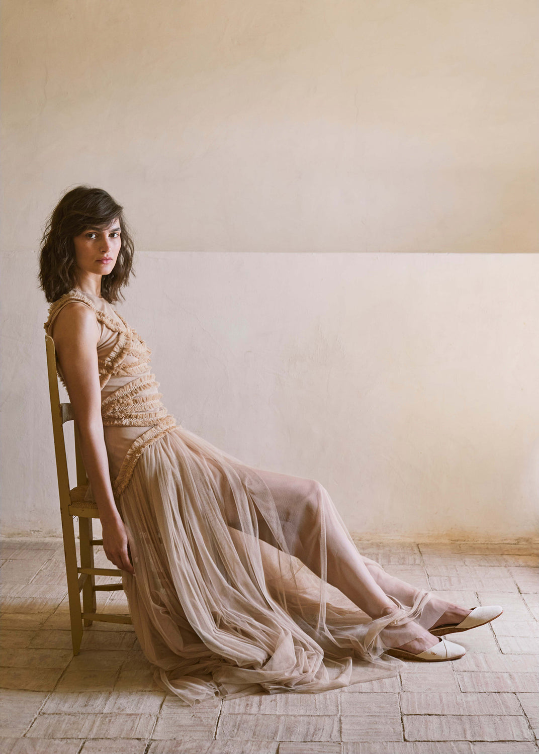Woman seated indoors wearing beige pointed-toe espadrille flats styled with a flowing neutral tulle dress in a warm Mediterranean interior.