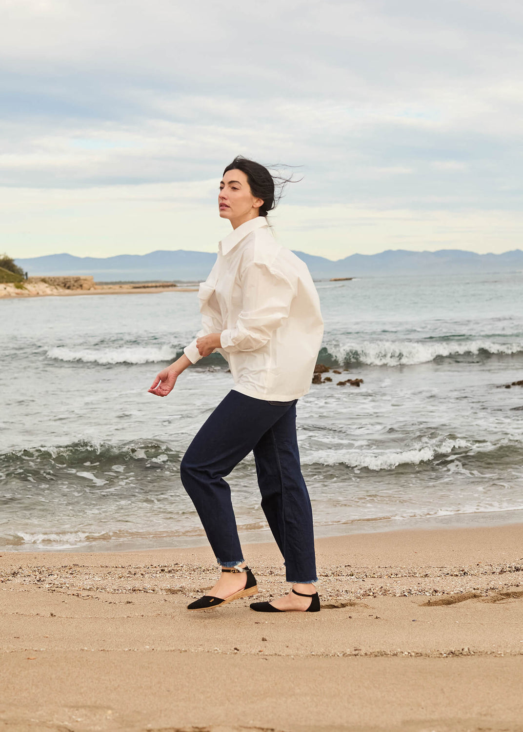 Woman walking along the shoreline in Montroig black canvas ankle-strap flat espadrilles, wearing a white shirt and navy trousers against a calm coastal backdrop.