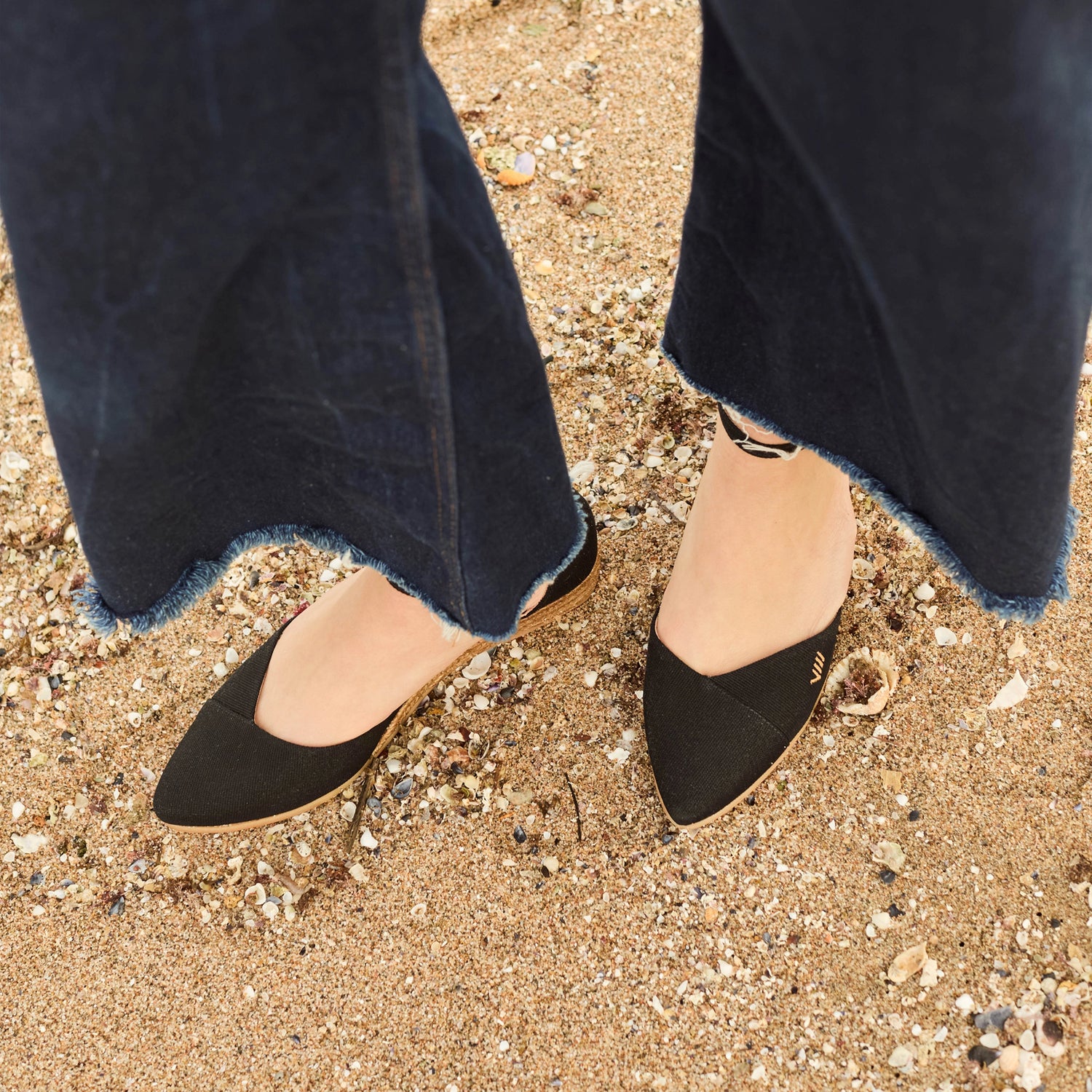 Top view of black espadrille ankle strap flats on sand, styled with dark denim
