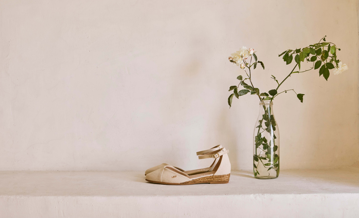Ivory beige ankle-strap espadrille flats styled beside fresh flowers in a minimalist Mediterranean interior.
