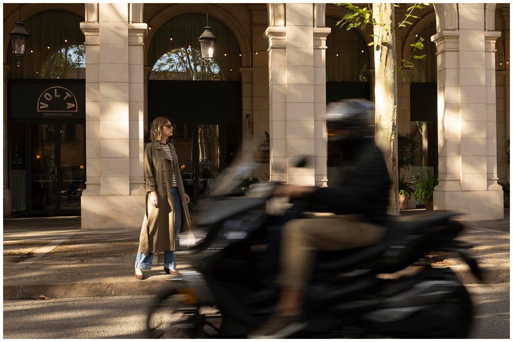 A woman in a long beige trench coat and blue jeans stands on a city sidewalk in front of a building with large stone columns, while a blurred motorcyclist passes in the foreground.