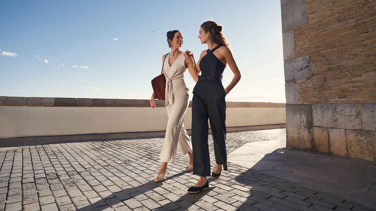 Two women walking in Sitges, one wearing a beige outfit paired by Sacova canvas beige peep open toe medium wedge espadrilles and the other in a black outfit paired with Garvet black suede flexible flat shoes
