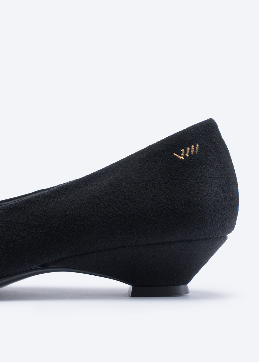 A close-up side view of a black wedge shoe with a textured suede-like finish and a small gold chevron logo on the heel, set against a plain white background.