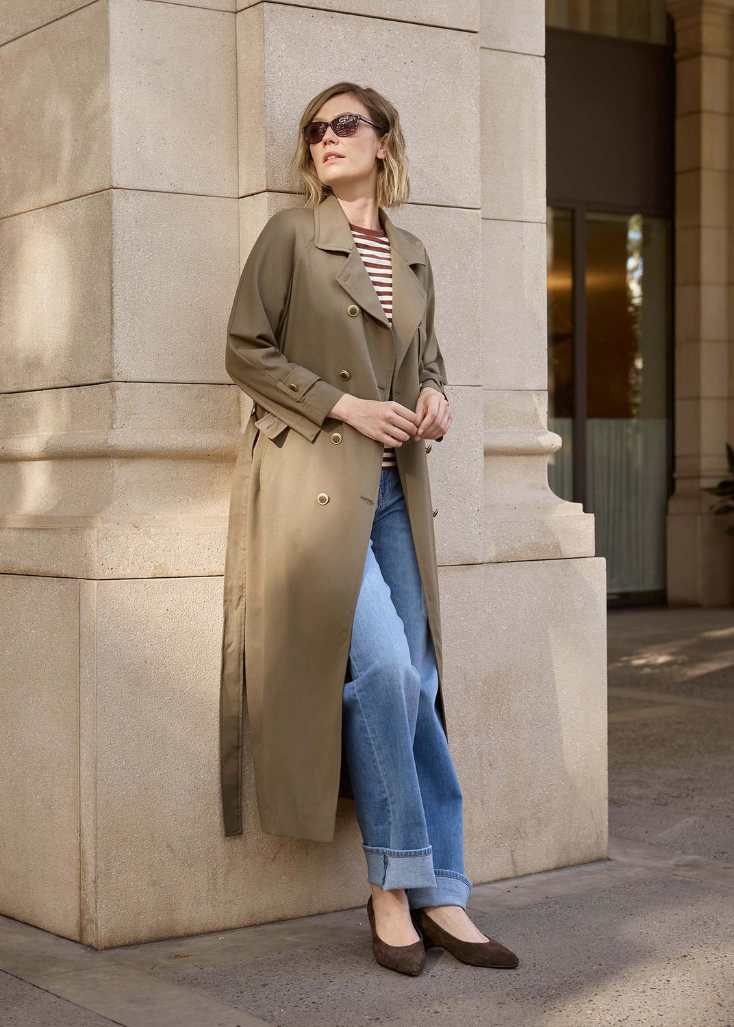 A woman wearing a long olive-brown trench coat over a striped top and blue jeans, standing outdoors against a beige stone building corner.