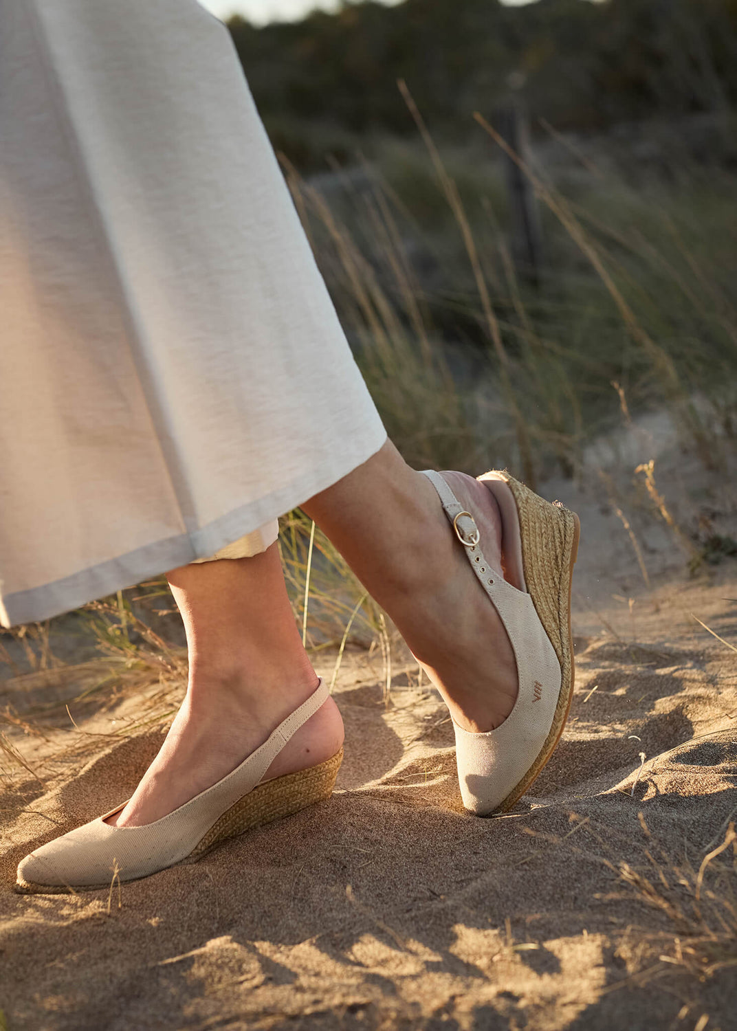 Close-up of a woman wearing Palomera beige canvas slingback wedge espadrilles, walking on sandy dunes at golden hour, capturing relaxed Mediterranean sophistication.