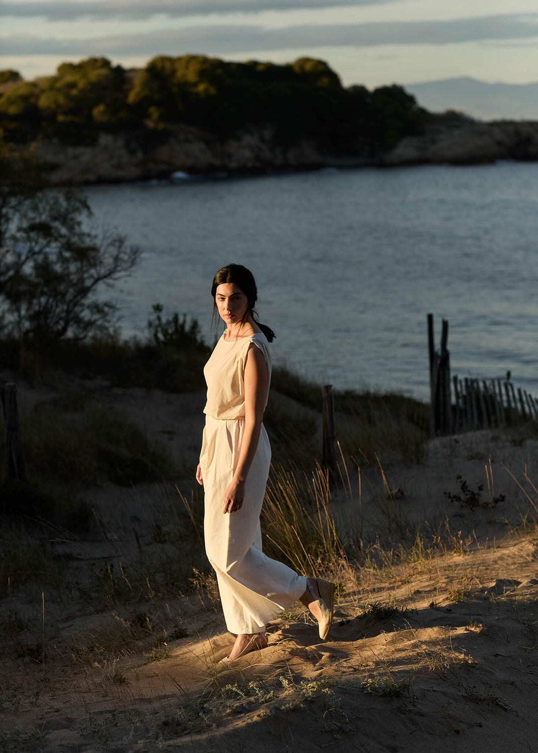 Woman walking along a sandy coastal path at sunset, wearing a light cream jumpsuit and Palomera beige canvas slingback wedge espadrilles.