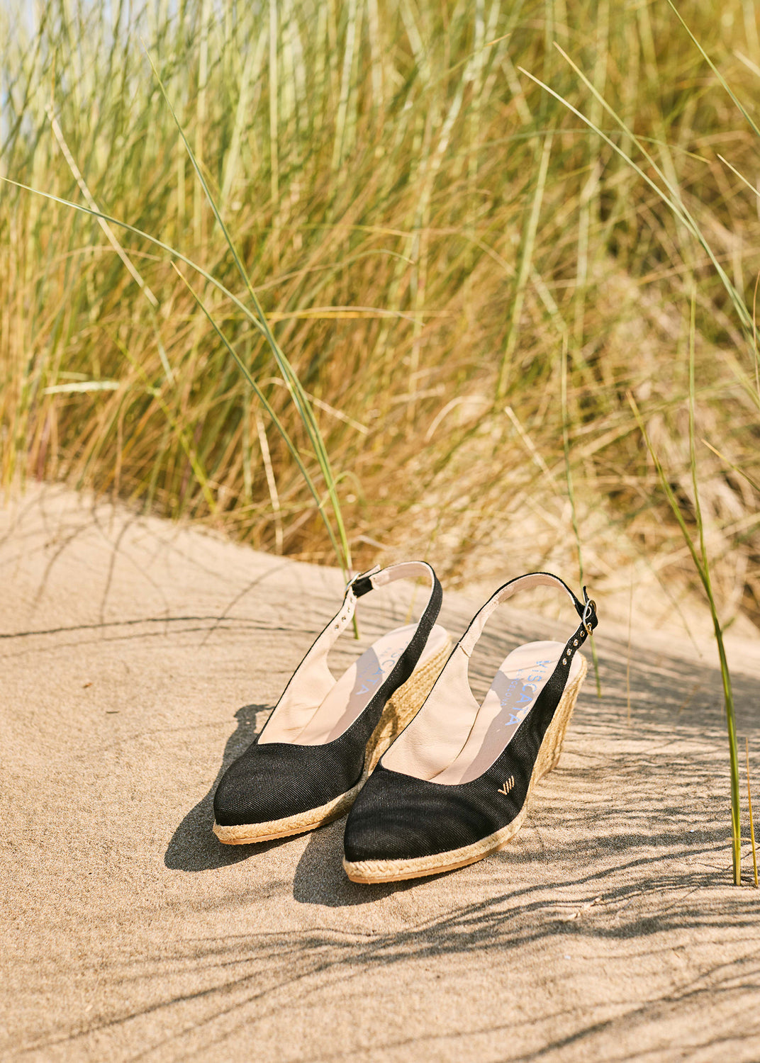 Black slingback espadrille wedges with braided jute soles placed on Mediterranean sand dunes in warm natural light.