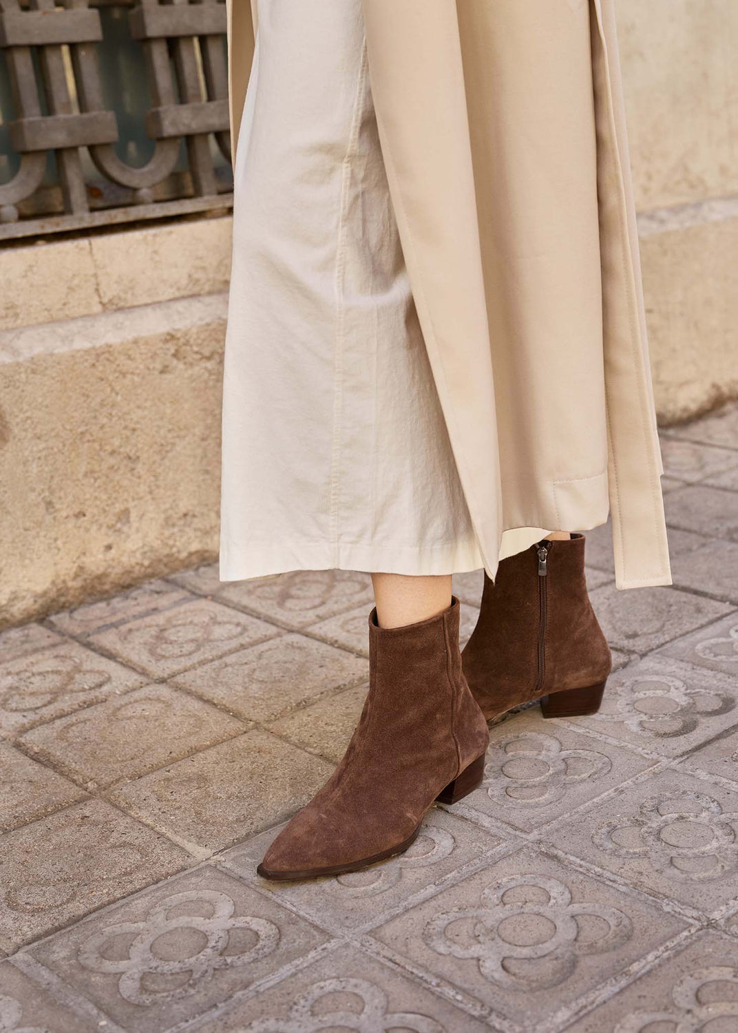 A person wearing brown suede ankle boots with low block heels, paired with a long beige skirt, standing on a paved stone street.