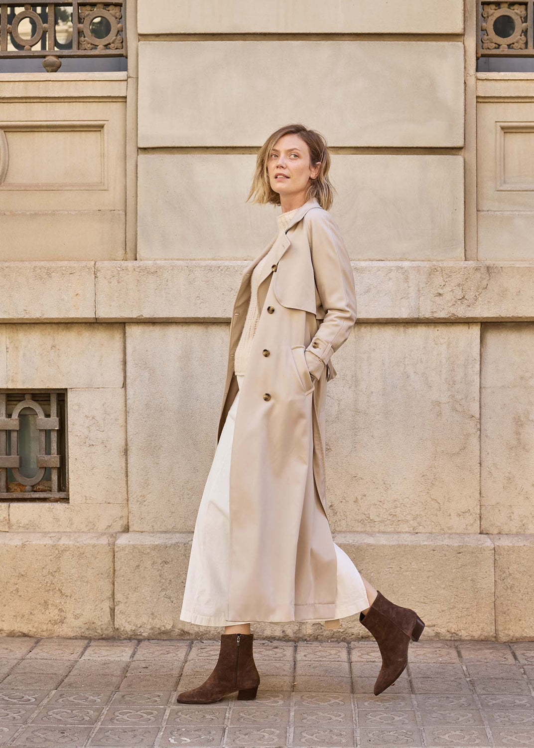A woman wearing a long beige trench coat and brown ankle boots walks along a stone sidewalk in front of a beige stone building.
