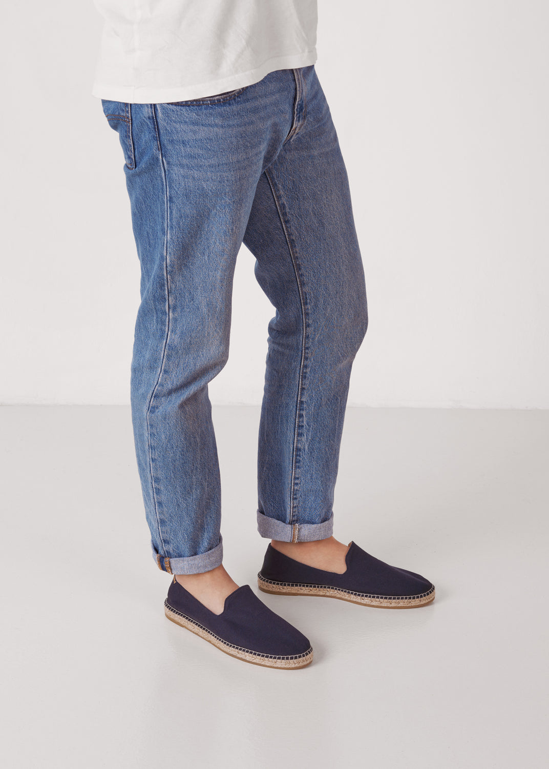 Men’s navy canvas slip-on espadrilles styled with cuffed jeans, photographed in a clean studio setting with the jute sole in view.